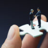 Hand holding puzzle piece with two small male figures on top shaking hands