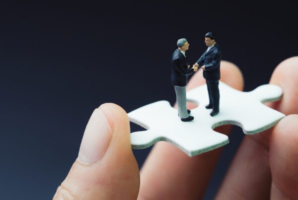 Hand holding puzzle piece with two small male figures on top shaking hands