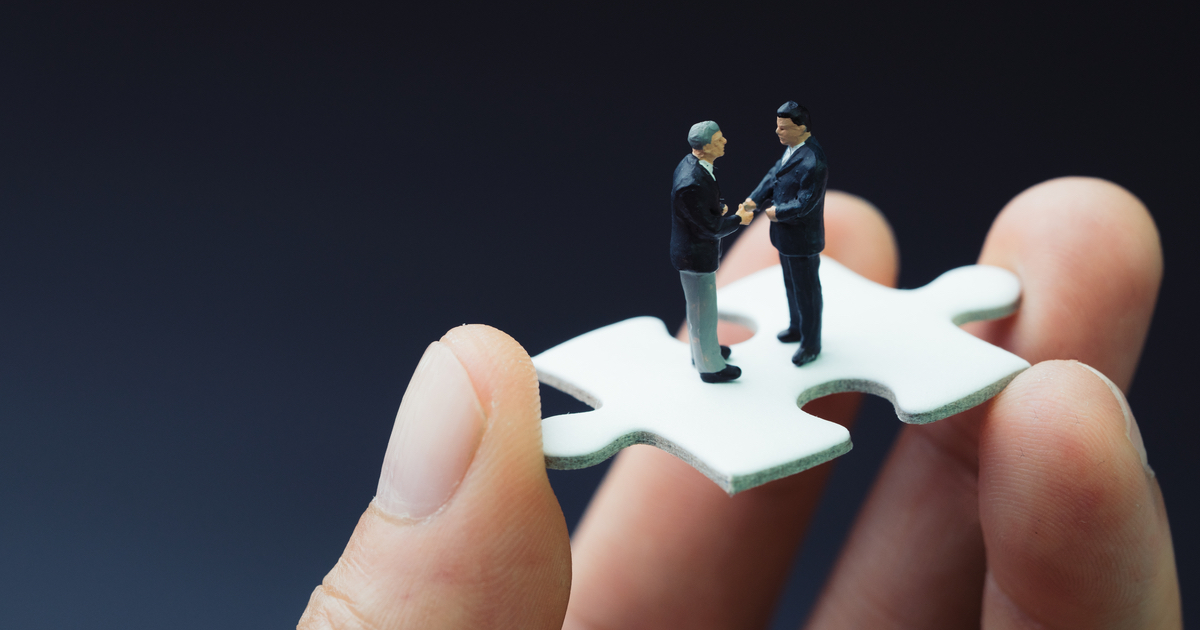 Hand holding puzzle piece with two small male figures on top shaking hands